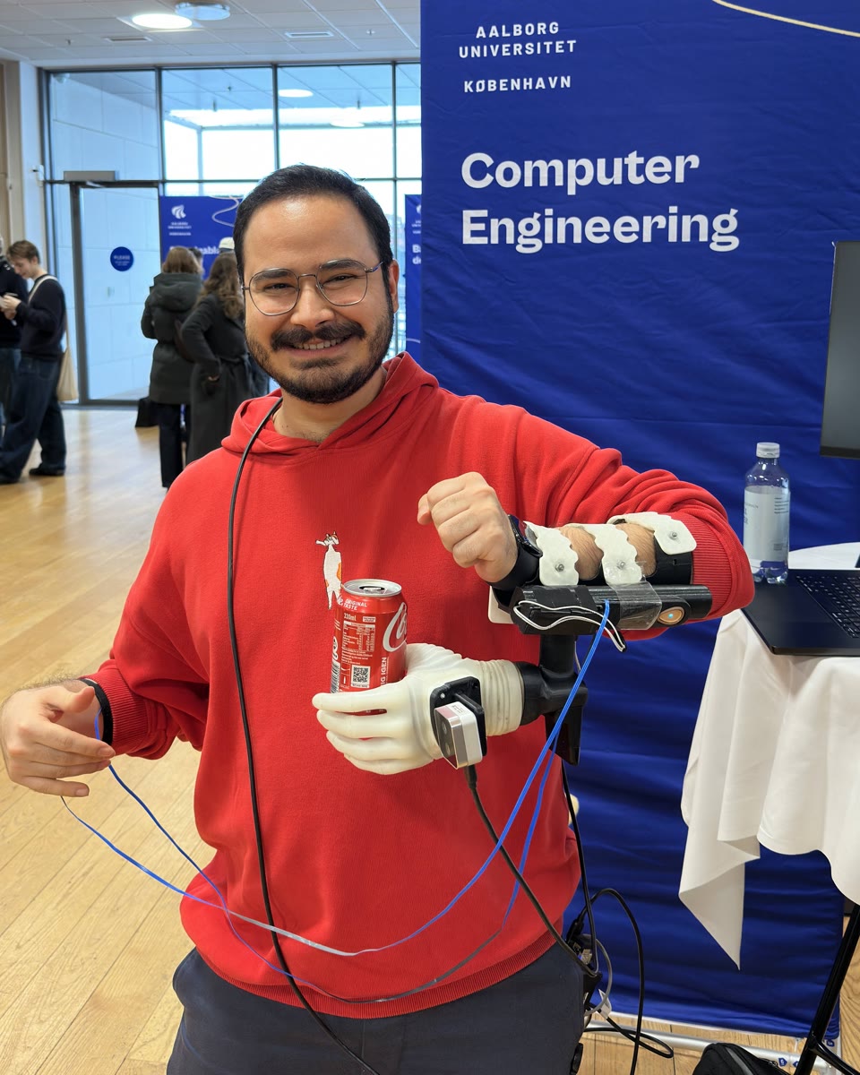 Ozan demonstrates a forearm-mounted robotic hand holding a Coca-Cola can at Aalborg University's Computer Engineering stand